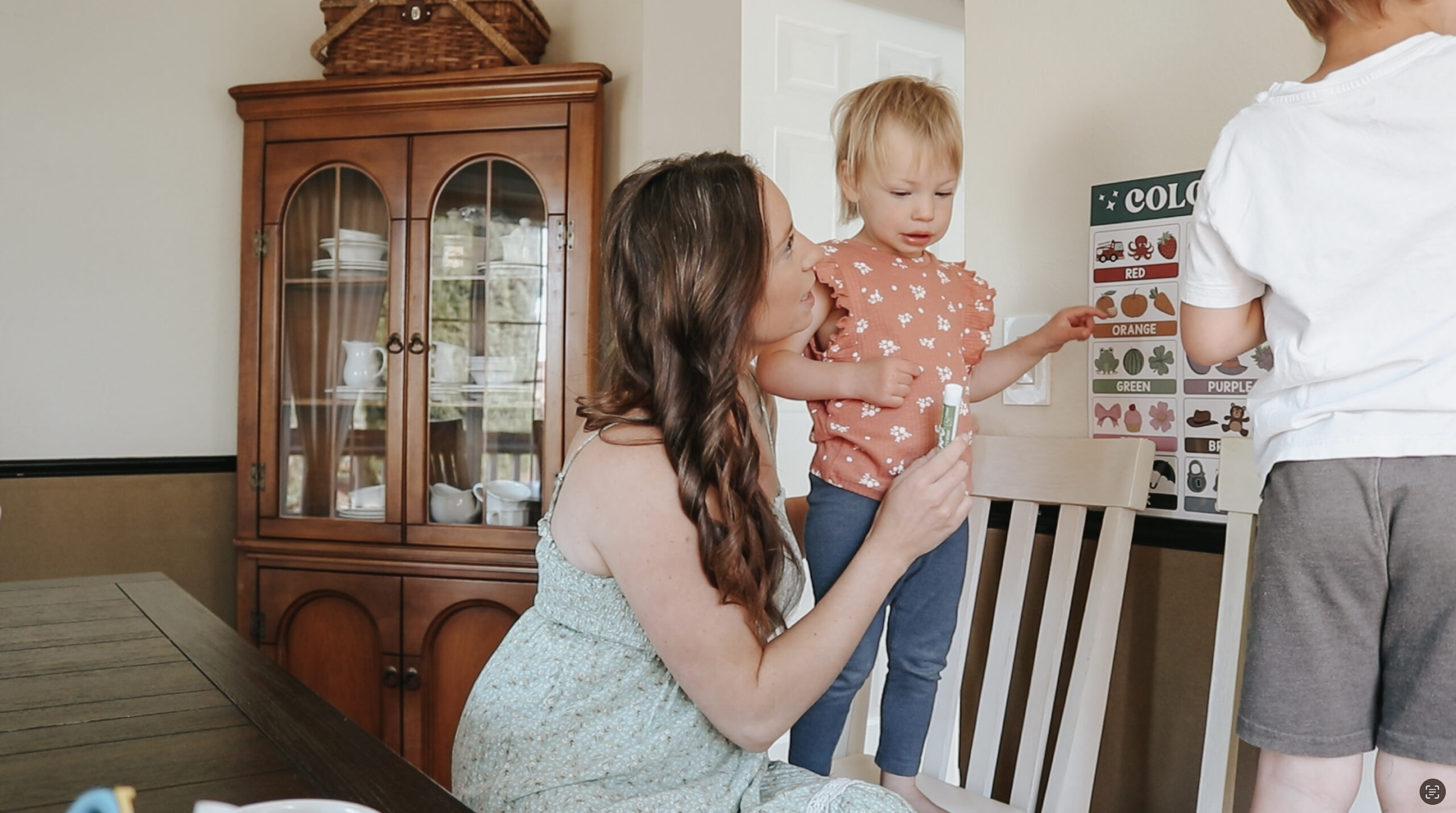 Christian homemaker sits at dining room table with her toddler daughter standing on her lap. There is a large china cabinet behind her.